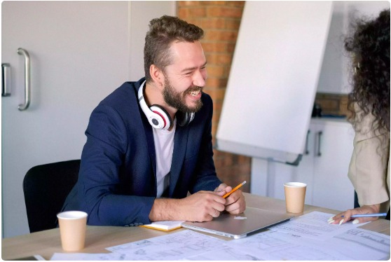 Homme souriant portant des écouteurs à un bureau avec un ordinateur portable et des tasses de café, engagé dans une discussion sur les services d'hébergement radio en ligne.