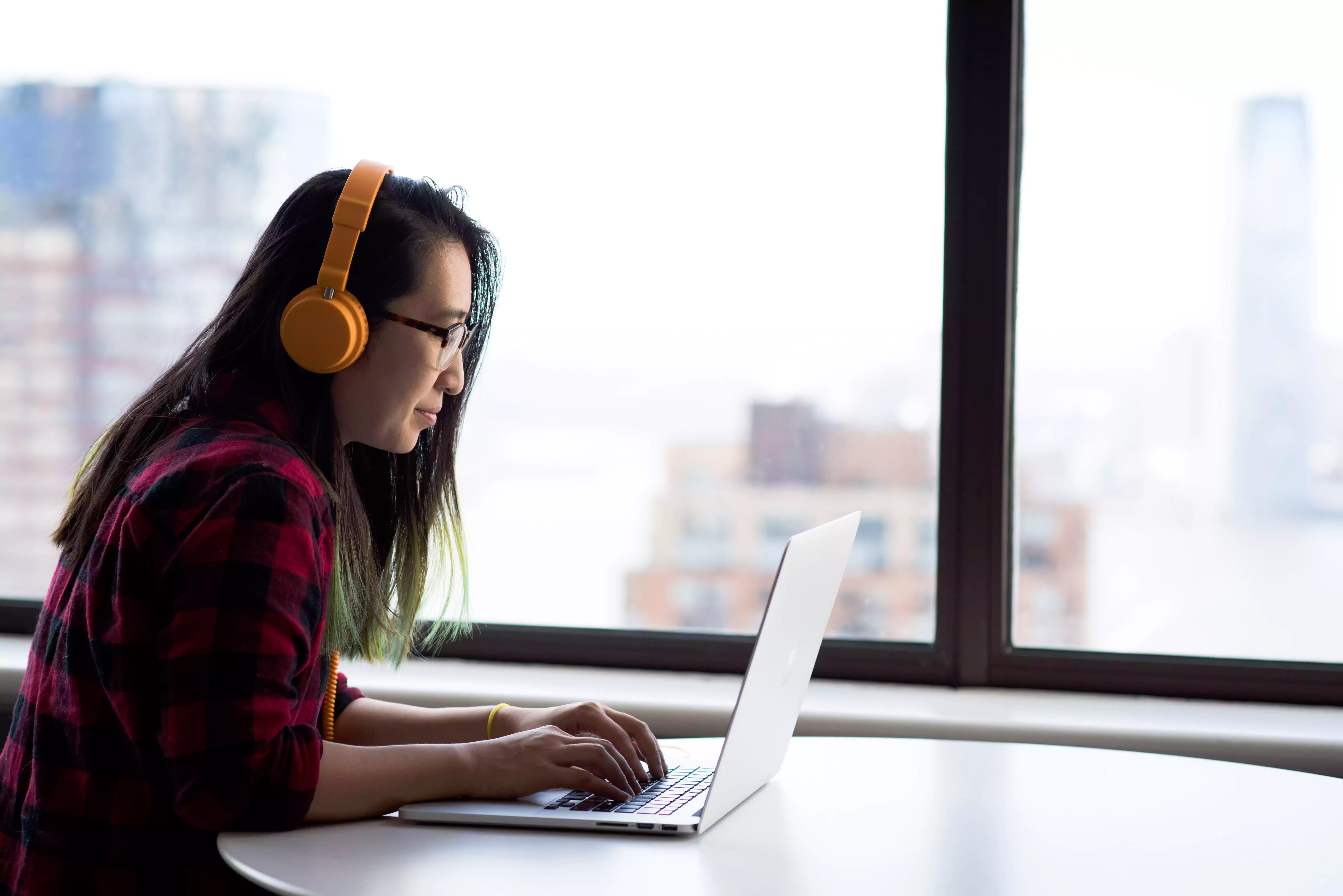 Person wearing orange headphones working on a laptop at a table by a window, illustrating remote work and online management for web hosting services.