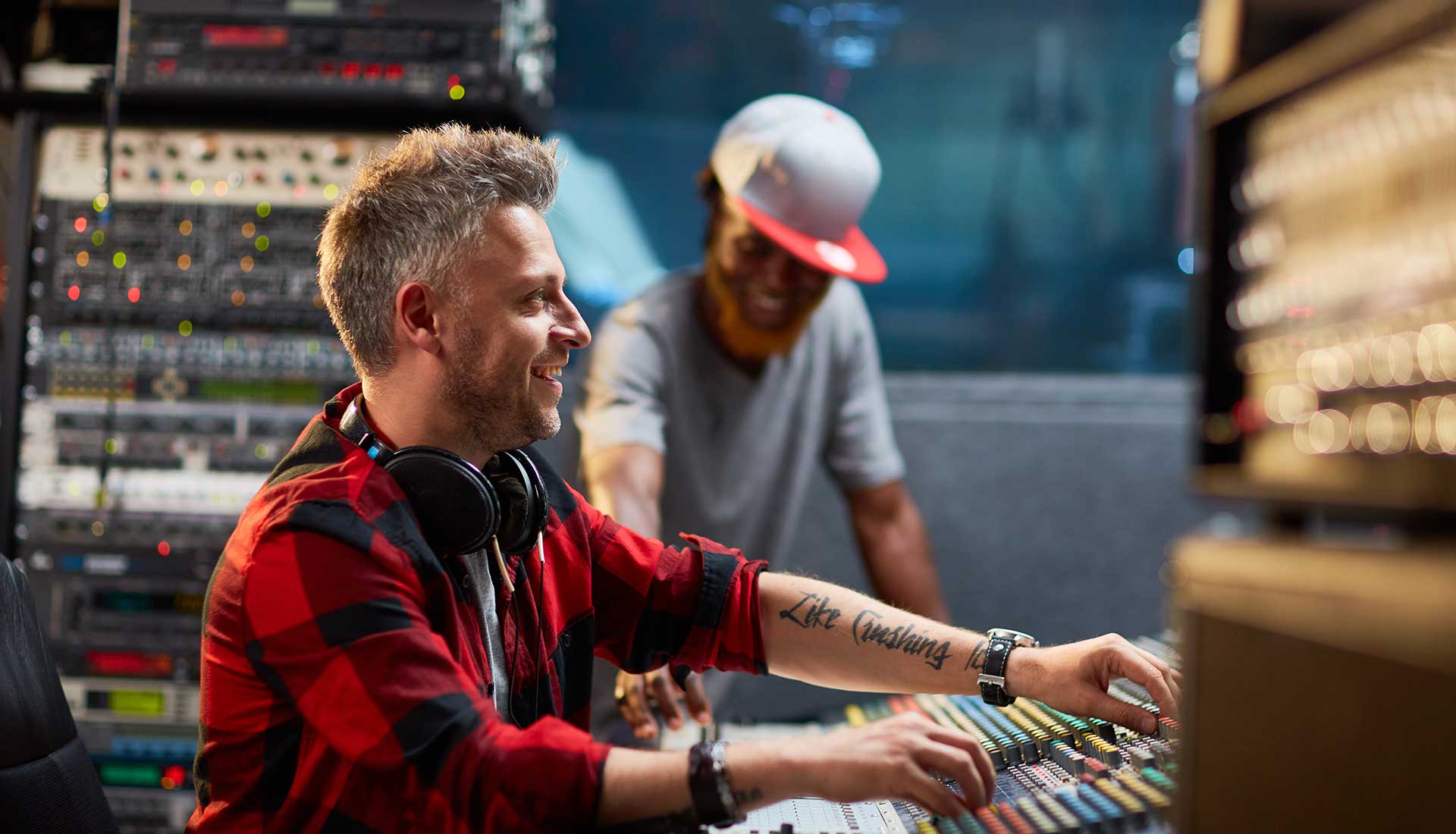 Two men in a radio studioone adjusting audio-mixer controls while wearing headphones and a red plaid shirt, the other smiling in the backgroundemphasizing CastHosts secure radio-streaming services.